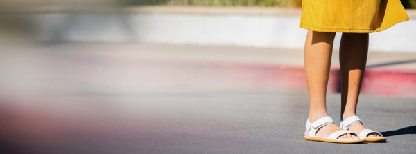 Pohľad na ženské nohy v bielych mestských sandáloch naboso, žena má na sebe horčicovo žltú sukňu.