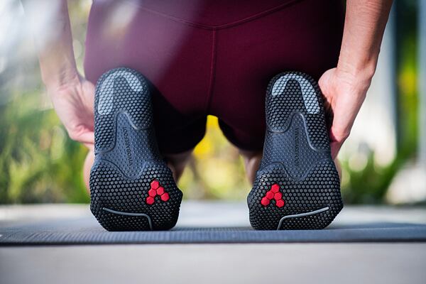 Žena sedí na podpätkoch, v detaile vidno podrážky bosých topánok. Topánky sú čierne s červeným logom V ako VIVOBAREFOOT.